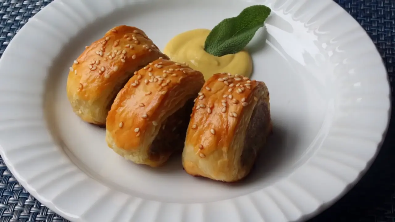 Afbeelding van een goudbruin worstenbroodje op een wit bord, gegarneerd met sesamzaadjes.